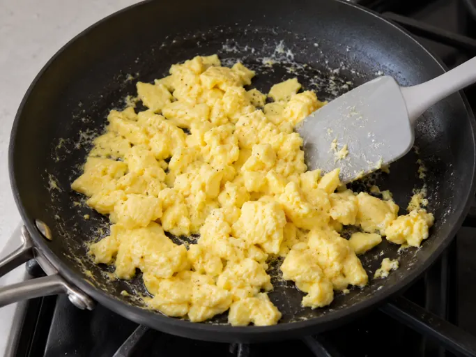 Illustrative step 1 for Buttery vegetable egg fried rice: Show three beaten eggs cooking as pale yellow, tender fully opaque and fully set scrambled curds in butter in a skillet, a plain spatula folding them